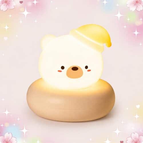 Une Veilleuse Kawaii en ours diffusant une lueur douce en forme d’ours blanc avec un petit bonnet jaune, posée sur une base ronde beige, sur fond rose pastel avec étoiles et cœurs.