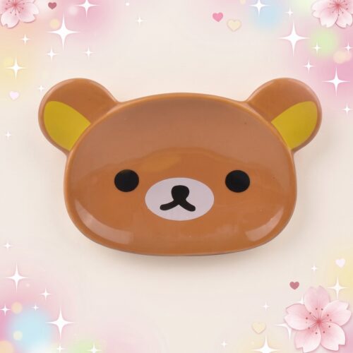 Un vaisselle kawaii plat à sauce ours mignon en forme de tête d’ours brun, avec de petites oreilles jaunes et un visage adorable, posé sur un fond pastel scintillant.