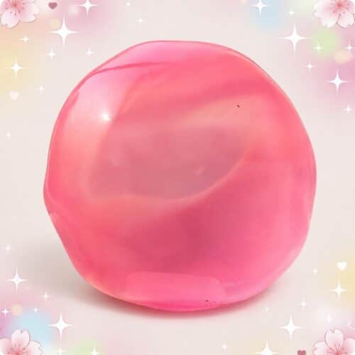 Une balle mochi squishy anti-stress rose brillante, ronde et moelleuse, posée sur un fond pastel avec des étoiles et des petits cœurs.