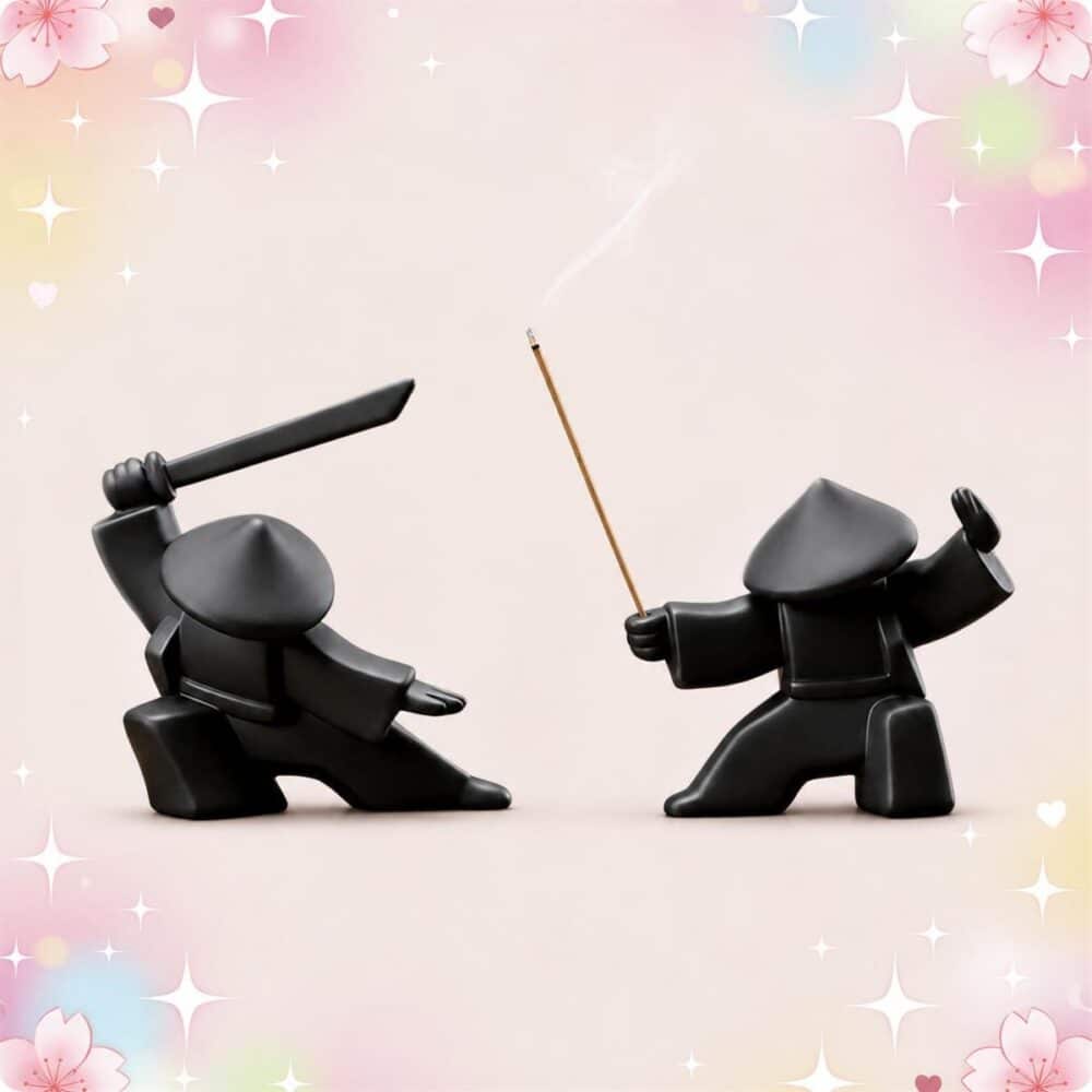 Deux figurines noires de samouraï pour décoration japonaise 2 brûleurs d'encens samouraï, l’une brandissant un sabre et l’autre tenant un bâton d’encens, sur fond rose doux.