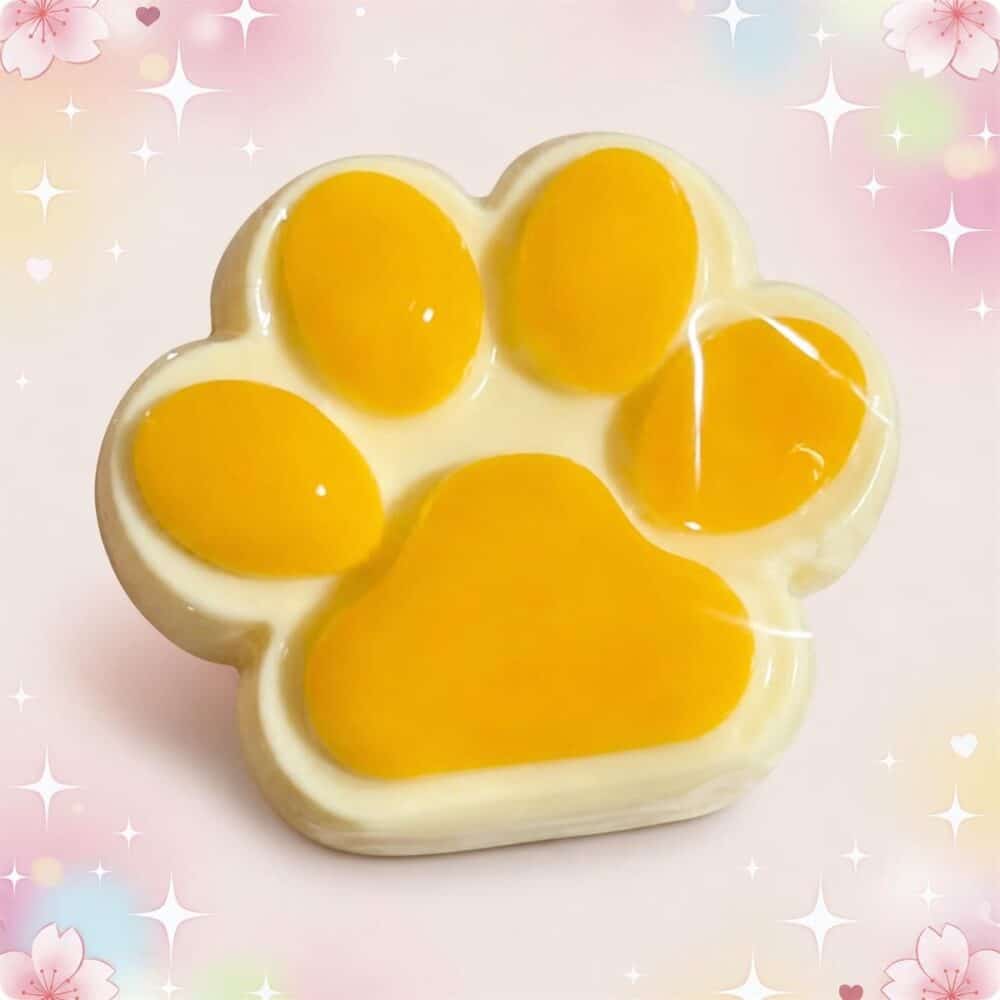 Un jouet anti-stress squishy en forme de patte de chat jaune et blanc, posé sur un fond rose pastel décoré de petites étoiles et de cœurs.
