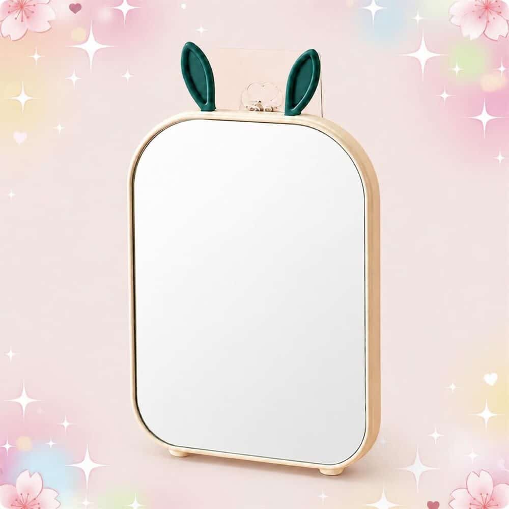 Un miroir Kawaii au design oreilles de lapin pliable, posé debout avec un cadre beige et deux petites oreilles vertes, sur un fond rose doux et scintillant.