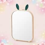 Un miroir Kawaii au design oreilles de lapin pliable, posé debout avec un cadre beige et deux petites oreilles vertes, sur un fond rose doux et scintillant.