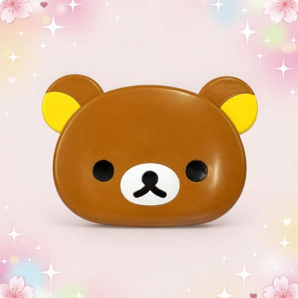 Un miroir kawaii Rilakkuma pliable avec peigne marron et jaune, en forme de tête d’ours, posé sur un fond rose doux et brillant.