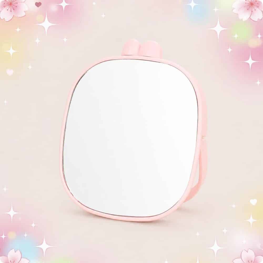 Un miroir kawaii rose portable à oreilles de lapin, avec une forme arrondie et un cadre rose, posé sur un fond pastel décoré d’étoiles et de petites fleurs.