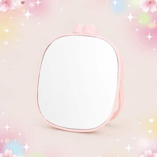Un miroir kawaii rose portable à oreilles de lapin, avec une forme arrondie et un cadre rose, posé sur un fond pastel décoré d’étoiles et de petites fleurs.