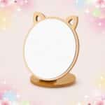 Un miroir de maquillage en bois pliable et rotatif avec oreilles de chat, posé sur un socle rond en bois, sur un fond rose doux et décoré d’étoiles scintillantes.
