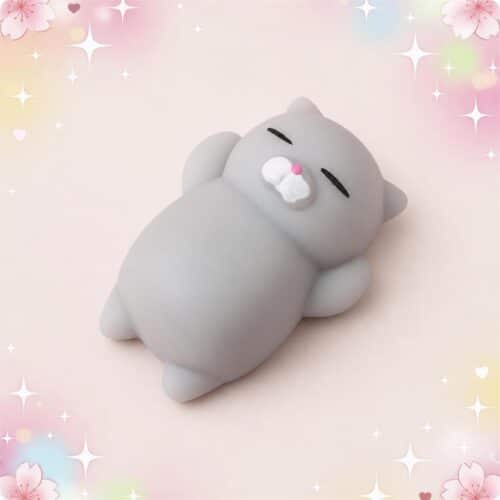 Un mochi squishy anti-stress en forme de chat gris, tout mignon et moelleux, posé sur un fond pastel avec des étoiles et des fleurs.
