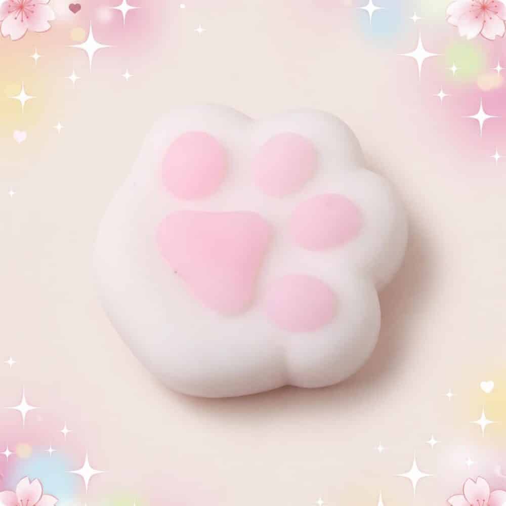 Un mochi squishy anti-stress en forme de patte de chat blanc et rose, tout doux à presser, sur un fond pastel décoré d’étoiles brillantes.