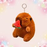 Un porte-clef peluche capybara mangeant une pastèque, doux et marron, suspendu à un anneau métallique sur fond rose avec des étoiles brillantes.