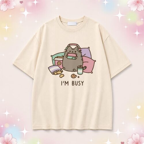 Un t-shirt kawaii Pusheen I'm Busy beige avec un chat gris mignon allongé sur des coussins pastel, sur fond rose doux avec des étoiles et des cœurs.