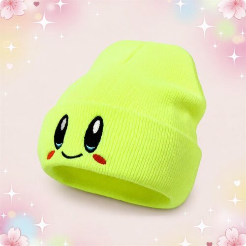 Un bonnet kawaii fluo à visage souriant brodé vert fluo, avec de grands yeux noirs et un petit sourire, posé sur un fond rose décoré d’étoiles et de cœurs.