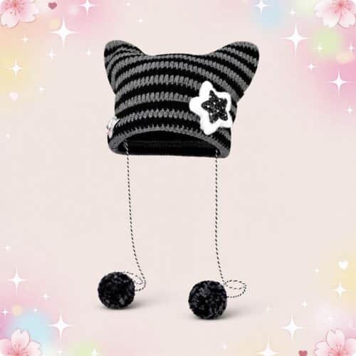 Un bonnet kawaii oreilles de chat rayé à pompons noir et gris, avec deux oreilles pointues, un badge étoile et deux cordons finis par des pompons, sur fond rose décoré de petites étoiles.