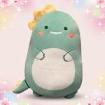 Un oreiller peluche dinosaure vert en forme de petit dinosaure souriant, avec ventre blanc, joues roses et fond pastel scintillant.