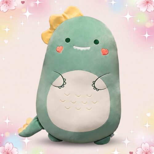 Un oreiller peluche dinosaure vert en forme de petit dinosaure souriant, avec ventre blanc, joues roses et fond pastel scintillant.