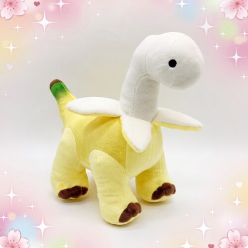 Une peluche dinosaure banane jaune et blanche, avec une tête de dinosaure drôle et des petites pattes marron, posée sur un fond pastel décoré d’étoiles et de cœurs.