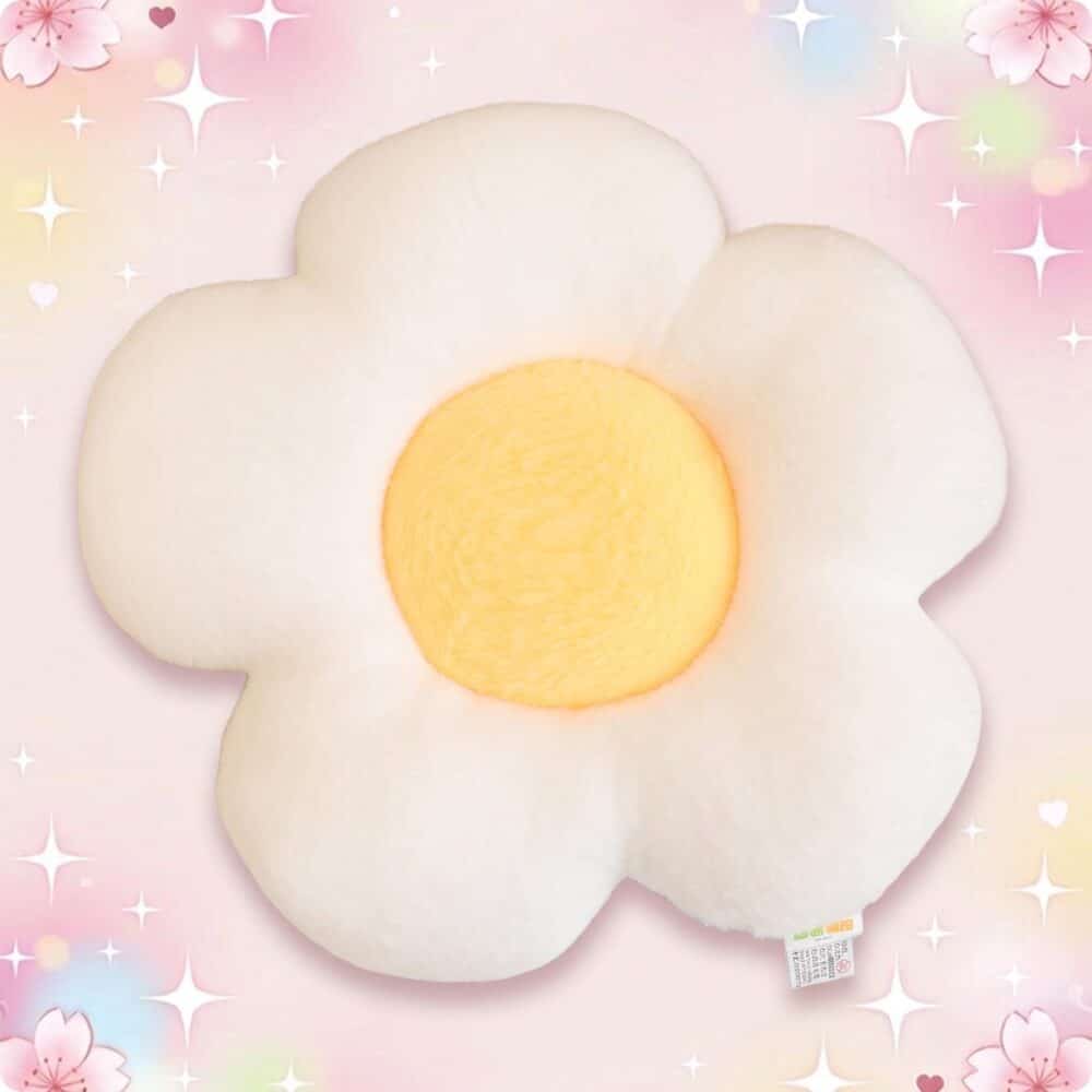 Un coussin kawaii marguerite tout doux, en forme de fleur blanche avec un centre jaune, posé sur un fond rose pastel avec des étoiles et des petites fleurs.