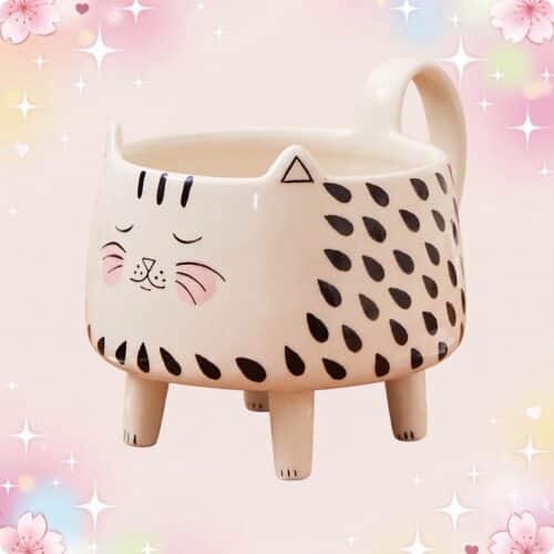 Une tasse kawaii chat sur pieds en céramique, avec un visage de chat souriant, des taches noires et de petites pattes, sur un fond rose et brillant.