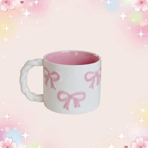 Une tasse kawaii à noeuds roses et anse torsadée blanche, avec intérieur rose, posée sur un fond pastel brillant avec des petits cœurs et étoiles.