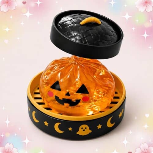 Un mochi squishy Halloween citrouille orange translucide et souriante, posé dans une boîte noire décorée de lunes et de petits fantômes, sur fond pastel brillant.
