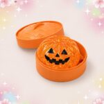 Un mochi squishy Halloween citrouille orange au visage sculpté, posé dans une petite boîte ronde orange, sur un fond clair décoré d’étoiles et de fleurs pastel.