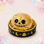 Un mochi squishy Halloween crâne doré à la bouche cousue, en forme de petit crâne brillant posé sur un support noir et jaune décoré de lunes et d’étoiles.