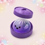 Un mochi squishy Halloween fantôme violet au regard fâché dans une boîte ronde violette, avec un petit fantôme blanc sur le côté, sur fond pastel brillant.