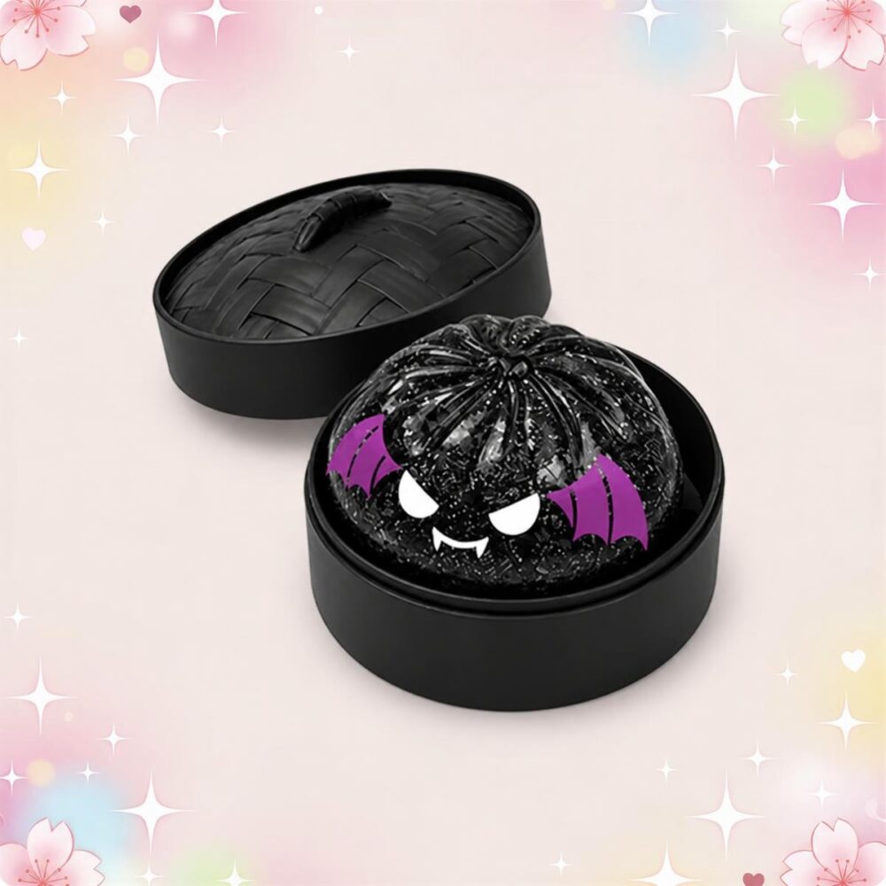 Un mochi squishy Halloween monstre noir aux ailes violettes présenté dans une boîte ronde noire, avec un visage de petit monstre et un fond rose décoré d’étoiles.
