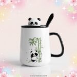 Un mug kawaii panda mignon avec couvercle blanc et noir, décoré d’un petit panda et de bambous, avec une cuillère et un couvercle en forme de tête de panda sur fond rose scintillant.