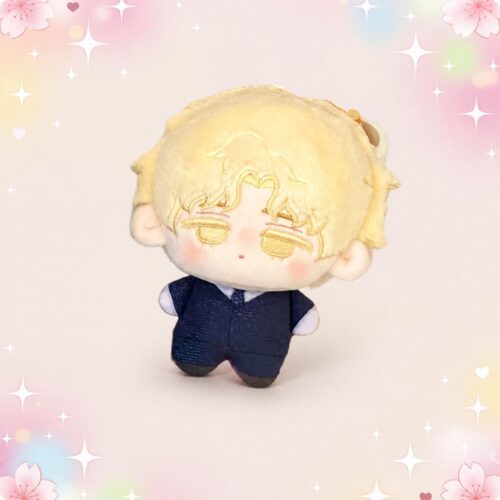 Une petite peluche aux cheveux blonds, aux joues roses et vêtue d’un costume bleu, représentant une mini peluche Alien Stage Luka, sur un fond pastel décoré de fleurs, de cœurs et d’étoiles.