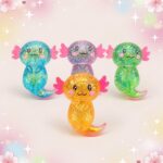 Quatre axolotls scintillants aux couleurs variées, avec des queues roses, présentent l’anti-stress squishy en axolotls scintillants aux queues roses sur un fond pastel décoré de fleurs et d’étoiles.