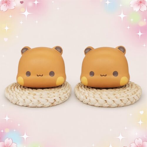 Deux mochi squishies orange en forme d’oursons souriants du lot de 2 mochi squishies Dudu ourson souriant, posés sur des dessous de verre tressés, sur un fond pastel décoré d’étoiles et de fleurs.
