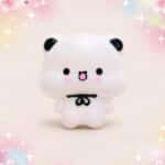 Un mochi squishy Bubu blanc avec un nœud papillon noir, issu du lot de 2 mochi squishies Bubu au nœud papillon, sur un fond rose décoré de fleurs et d’étoiles.