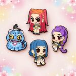 Un lot de 4 pins chibi Kpop Demon Hunters représentant trois personnages et un chat bleu, disposés sur un fond rose pastel décoré de fleurs et d’étoiles.